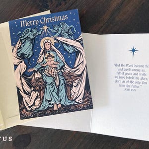 May include: A folded Christmas greeting card featuring a dark blue illustration of the Virgin Mary holding the infant Jesus, flanked by two angels under a star. The interior displays the text: And the Word became flesh and dwelt among us.