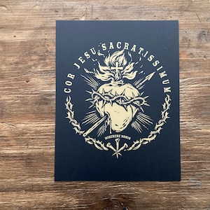 May include: A black and gold print of a stylized heart with a crown of thorns, flames, and a cross. The text around the heart reads "COR JESU SACRATISSIMUM". The text below the heart reads "MISERERE NOBIS".