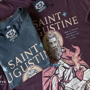 May include: Two t-shirts, one dark gray and one burgundy, with the text "SAINT AUGUSTINE" and an illustration of a saint. The shirts have tags that read "BARITUS CATHOLIC TEES". The burgundy shirt has the quote "...and our hearts are restless until they rest in You."