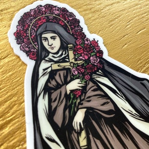 May include: A sticker featuring a saintly figure with a halo of pink roses, holding a bouquet of roses and a cross. The figure is dressed in a black veil and brown robes, with a white undergarment. The sticker has a white border.