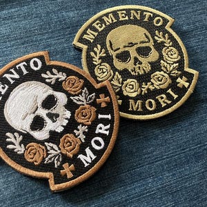May include: Two embroidered patches with a skull design and the words "Memento Mori". One patch features a white skull with tan roses, the other has a gold skull. Both are on a black background with tan trim.