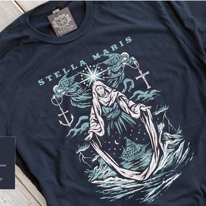 Op de afbeelding: Marineblau unisex T-shirt met korte mouwen en een grafisch ontwerp. Het ontwerp toont de woorden "STELLA MARIS" boven een illustratie van een religieuze figuur, engelen, een schip en een anker.