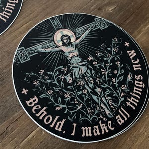 Può includere: Adesivo rotondo con sfondo nero raffigurante Gesù sulla croce, circondato da elementi floreali. Il testo "Ecco, io faccio nuove tutte le cose" è scritto attorno al bordo. L'adesivo ha un bordo bianco.