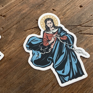 May include: A sticker featuring a religious image of a woman in a blue robe, holding a rope in her hands. The woman has a halo above her head and is depicted in a stylized, illustrative style.