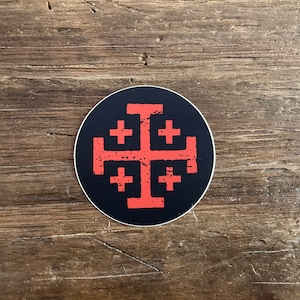 May include: A black and red round sticker with a red cross with four smaller crosses inside. The sticker has a distressed look.