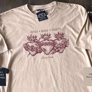 Könnte beinhalten: Cremefarbenes Kurzarm-T-Shirt mit einer Grafik von drei Herzen, jeweils mit unterschiedlichen Designs, und den Worten "Jesus + Mary + Joseph" und "Save Souls". Das Shirt hat ein Etikett mit der Aufschrift "Baritus Catholic Tees".