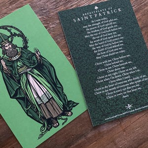 Op de afbeelding: Twee kaarten op een houten oppervlak. Eén kaart toont een illustratie van Sint Patrick in groene gewaden, met een staf. De andere kaart heeft de tekst "BREASTPLATE OF SAINT PATRICK" en een gebed. De kaarten zijn groen en wit.