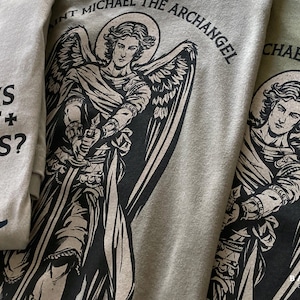 Puede incluir: Tres camisetas en beige y verde oliva con un estampado gráfico negro del Arcángel San Miguel con alas y una espada. El texto dice "SAINT MICHAEL THE ARCHANGEL" y "QUIS UT DEUS?" con pequeños símbolos de cruz en el diseño.