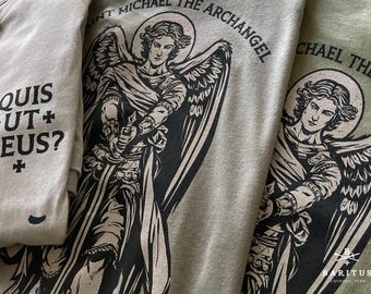 May include: Three t-shirts in beige and olive green featuring a black graphic print of Saint Michael the Archangel with wings and a sword. The text reads "SAINT MICHAEL THE ARCHANGEL" and "QUIS UT DEUS?" with small cross symbols included in the design.