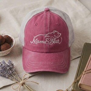 May include: A pink baseball cap with a white mesh back and a curved brim. The cap features the words "Mama Bear" in cursive with a bear graphic. The cap is sitting on a light-colored surface with a book, a small bowl, and a bouquet of lavender.