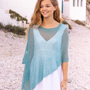 Könnte beinhalten: Ein hellblaues, asymmetrisches Strickoberteil mit V-Ausschnitt, getragen über einem weißen Kleid. Das Oberteil hat Dreiviertelärmel und eine transparente, offene Webart. Der Hintergrund zeigt ein weißes Gebäude und einen Kiesstrand.