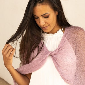 May include: A woman wearing a light pink knitted shawl over a white top. The shawl is loose and flowing, and the woman is looking down at her hands.