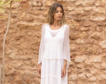 May include: A white maxi dress with a lace overlay and a tiered skirt. The dress is worn by a woman with long blonde hair, standing in front of a stone wall.