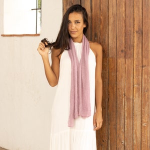 May include: A woman wearing a white maxi dress with a light purple scarf around her neck. She is standing in front of a wooden door.