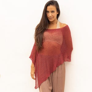 Puede incluir: Una parte superior asimétrica estilo poncho de color burdeos con un tejido abierto y transparente. La parte superior se combina con pantalones sueltos de color marrón claro. El conjunto se encuentra sobre un fondo blanco.
