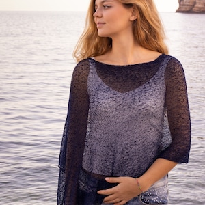May include: A navy blue knit poncho with a wide neckline and short sleeves. The poncho is layered over a light blue top and paired with denim shorts. The image is taken outdoors near the water.
