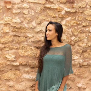 May include: A woman with long dark hair is wearing a teal green knitted poncho with a sheer panel in the front. She is standing in front of a stone wall.