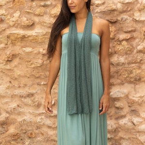 May include: A woman wearing a long, sage green dress with a matching green scarf. She is standing in front of a stone wall.