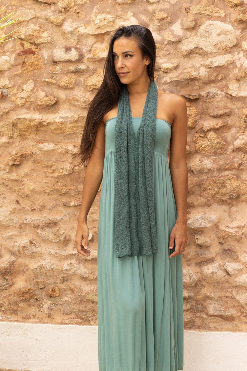 May include: A woman wearing a long, sage green dress with a matching green scarf. She is standing in front of a stone wall.