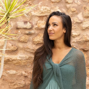 May include: A woman with long dark hair wearing a green knitted shawl stands in front of a stone wall.