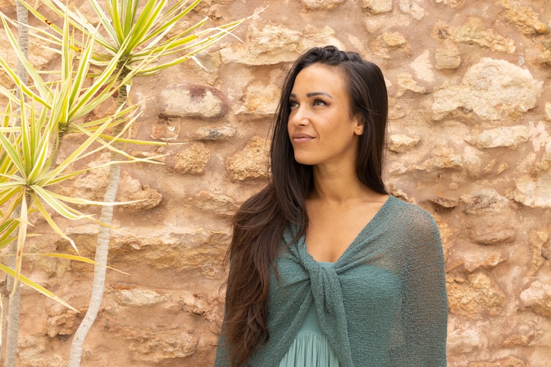 May include: A woman with long dark hair wearing a green knitted shawl stands in front of a stone wall.