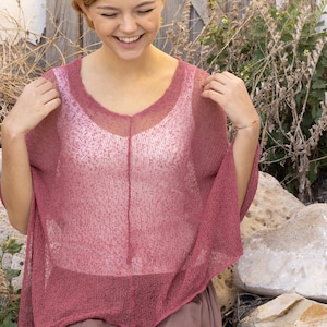May include: A woman wearing a loose-fitting, pink, crocheted top with a white tank top underneath. She is sitting on a stone step with a wooden door and plants in the background.