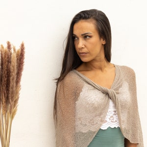 May include: A woman with long brown hair is wearing a light brown knitted shawl with a knotted front. The shawl is semi-transparent and shows a white lace top underneath. The woman is looking to the left of the frame.