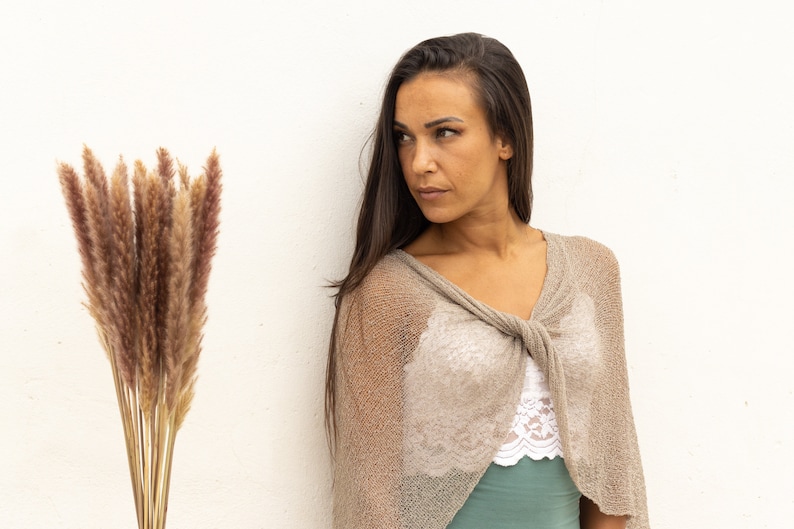May include: A woman with long brown hair is wearing a light brown knitted shawl with a knotted front. The shawl is semi-transparent and shows a white lace top underneath. The woman is looking to the left of the frame.