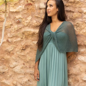 May include: A woman wearing a teal maxi dress with a matching teal crochet shawl tied around her neck. She is standing in front of a stone wall.