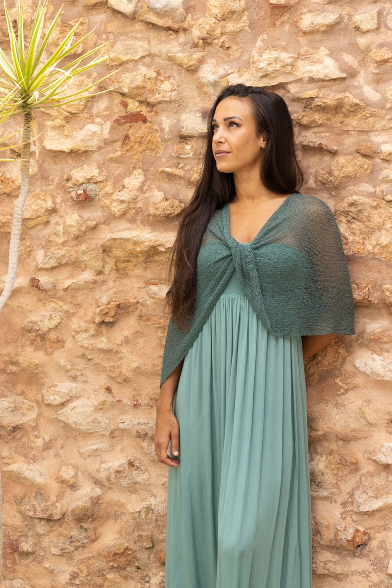 May include: A woman wearing a teal maxi dress with a matching teal crochet shawl tied around her neck. She is standing in front of a stone wall.