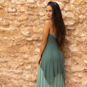 May include: A woman wearing a long, flowing, teal green dress with a layered skirt. She is standing in front of a stone wall.