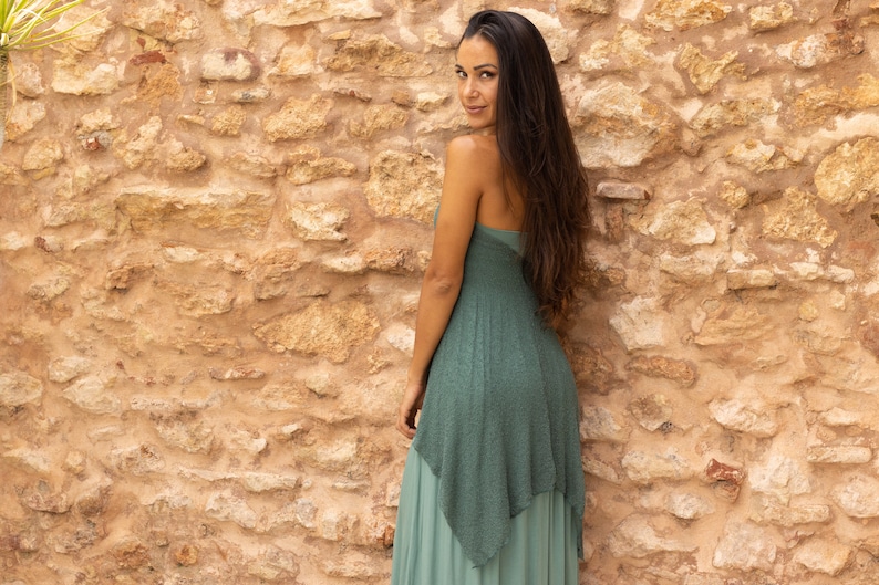 May include: A woman wearing a long, flowing, teal green dress with a layered skirt. She is standing in front of a stone wall.