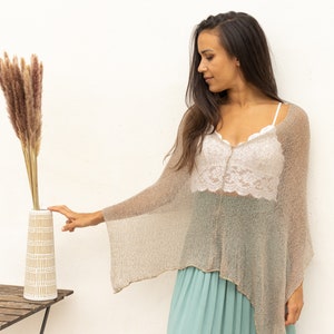 May include: A woman wearing a light brown knitted poncho and a white lace top, reaches out to touch a vase with dried pampas grass. The vase is white with a brown geometric pattern and is sitting on a wooden table.