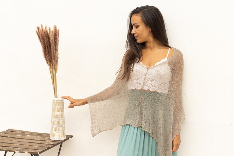 May include: A woman wearing a light brown knitted poncho and a white lace top, reaches out to touch a vase with dried pampas grass. The vase is white with a brown geometric pattern and is sitting on a wooden table.