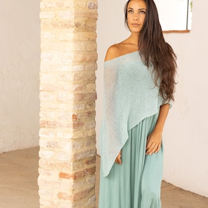May include: A woman wearing a light blue off-the-shoulder dress with a matching light blue knitted shawl. She is standing in front of a brick wall.