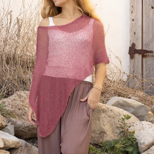May include: A woman wearing a loose-fitting, pink, knitted poncho with a long, brown skirt. She is standing in front of a white wall with a wooden door.