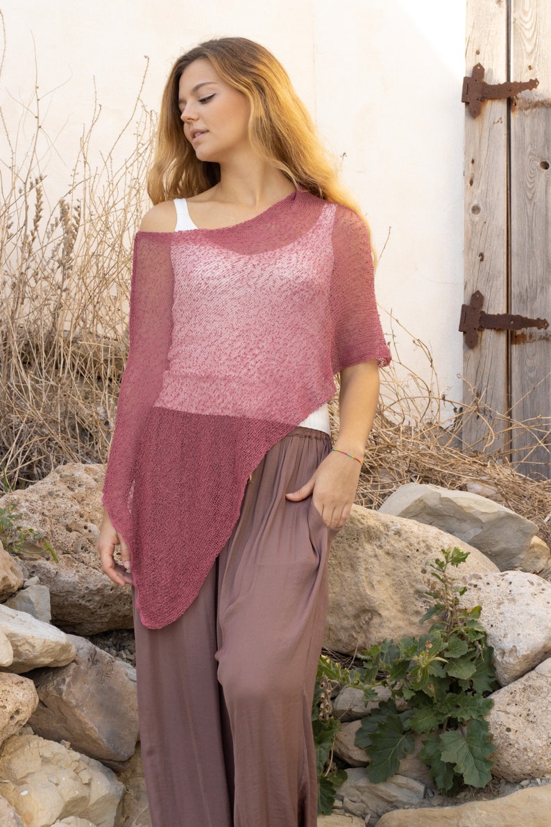 May include: A woman wearing a loose-fitting, pink, knitted poncho with a long, brown skirt. She is standing in front of a white wall with a wooden door.