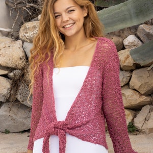 May include: A woman wearing a pink crocheted cardigan tied at the waist over a white tank top. She is standing in front of a stone wall with greenery.