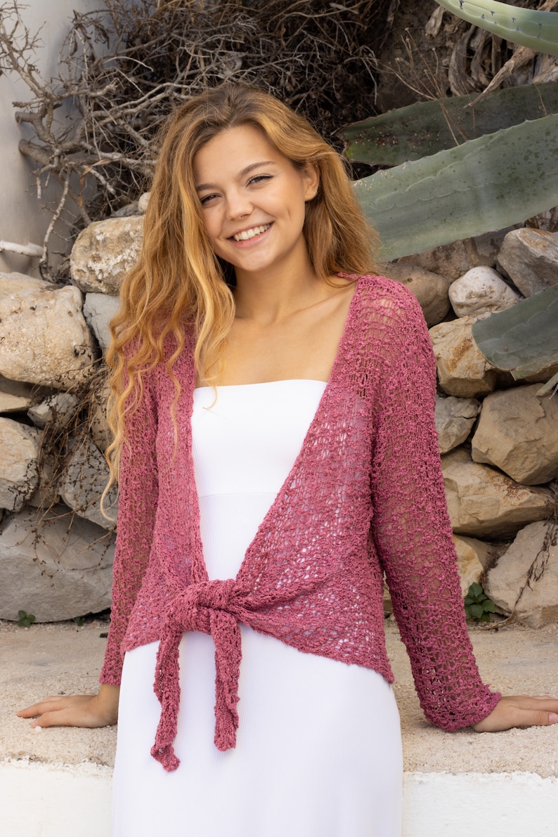 May include: A woman wearing a pink crocheted cardigan tied at the waist over a white tank top. She is standing in front of a stone wall with greenery.