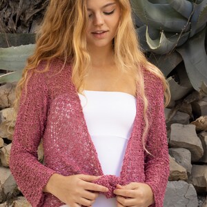 May include: A pink crocheted cardigan with a loose fit and a tie front. The cardigan is made of a lightweight, airy fabric and has a relaxed, bohemian style.