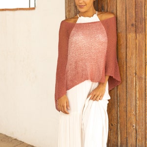 May include: A woman wearing a long white skirt and a loose-fitting, ombre pink knitted poncho. The poncho has a slightly scalloped hemline and is made of a soft, lightweight fabric.