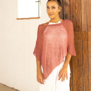 May include: A woman wearing a loose-fitting, dusty rose knitted poncho over a white maxi dress. She has her hair pulled back in a bun.