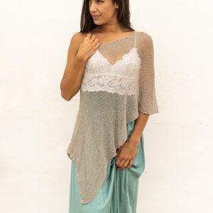 May include: A woman wearing a light brown, one-shoulder, knitted poncho over a white lace tank top and a teal maxi skirt.