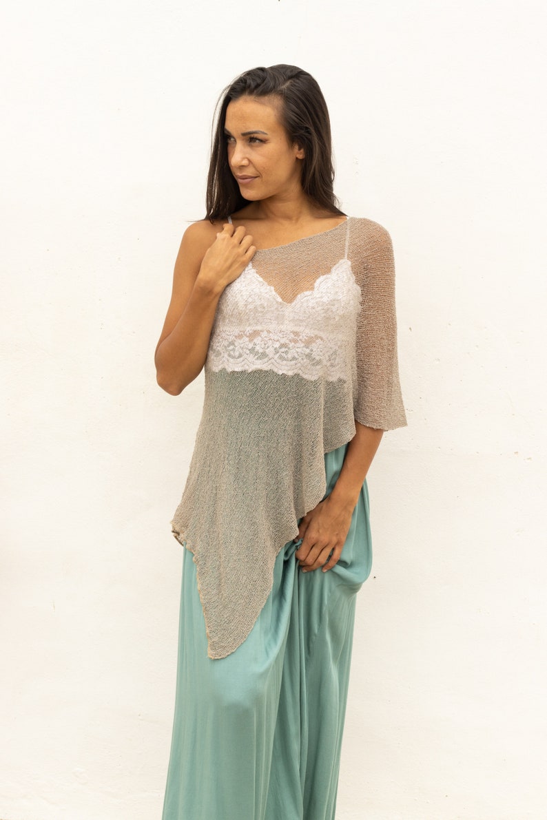 May include: A woman wearing a light brown, one-shoulder, knitted poncho over a white lace tank top and a teal maxi skirt.