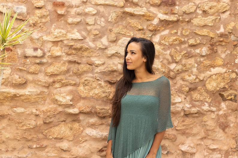 May include: A woman with long dark hair is wearing a teal green knitted poncho with a sheer panel in the front. She is standing in front of a stone wall.