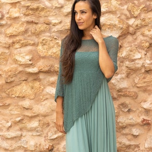 May include: A woman wearing a teal green knit top with a long teal green skirt. The top has a sheer panel in the front. She is standing in front of a stone wall.