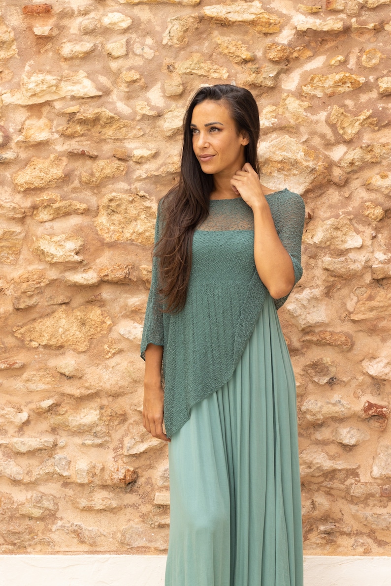 May include: A woman wearing a teal green knit top with a long teal green skirt. The top has a sheer panel in the front. She is standing in front of a stone wall.