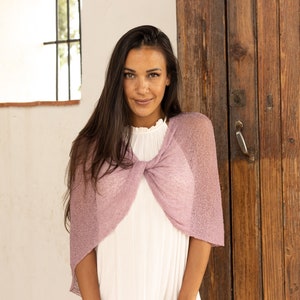 May include: A woman wearing a light pink knitted shawl over a white top. The shawl has a loose, open weave and is tied in a knot at the front.