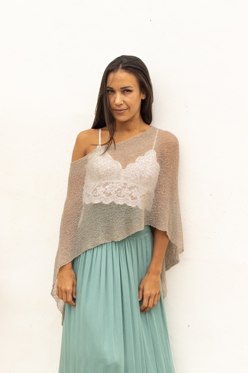 May include: A woman wearing a light brown crochet poncho over a white lace top and a teal maxi skirt.