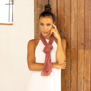 May include: A woman with brown hair pulled back in a bun is wearing a white sleeveless dress and a dusty rose colored scarf tied around her neck. She is standing in front of a wooden door with her hand on her chin.